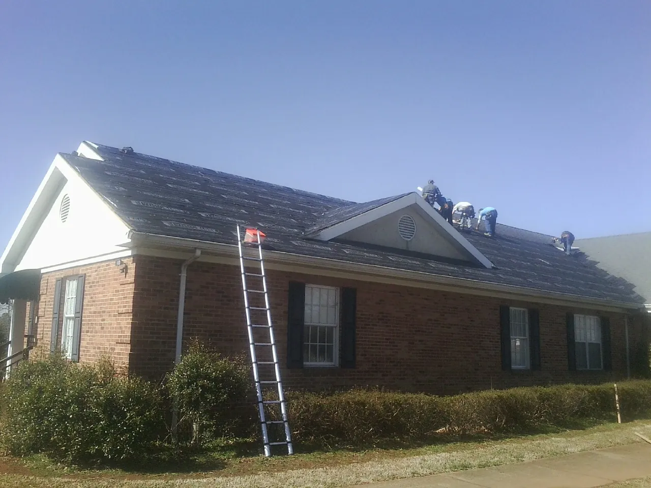 Crew tearing shingles off a brick ranch roof with a ladder set — Emergency Roof Repair work by our crew in Pinehurst